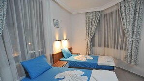 A hotel room with two beds, each with a blue bedspread and white towels. There are two wall-mounted lamps, a framed picture on the wall, and sheer curtains on the windows. - Happy Homes Apartments (Alanya)