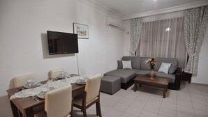 A modern living room with a dining area, a grey sofa, and a flat-screen TV mounted on the wall. - Happy Homes Apartments (Alanya)