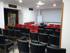 Un salón de conferencias con filas de sillas negras, una pizarra y una pantalla para proyector.
