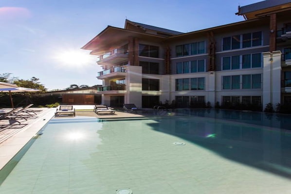 A modern hotel with a large swimming pool, multiple balconies, and a clear sky.