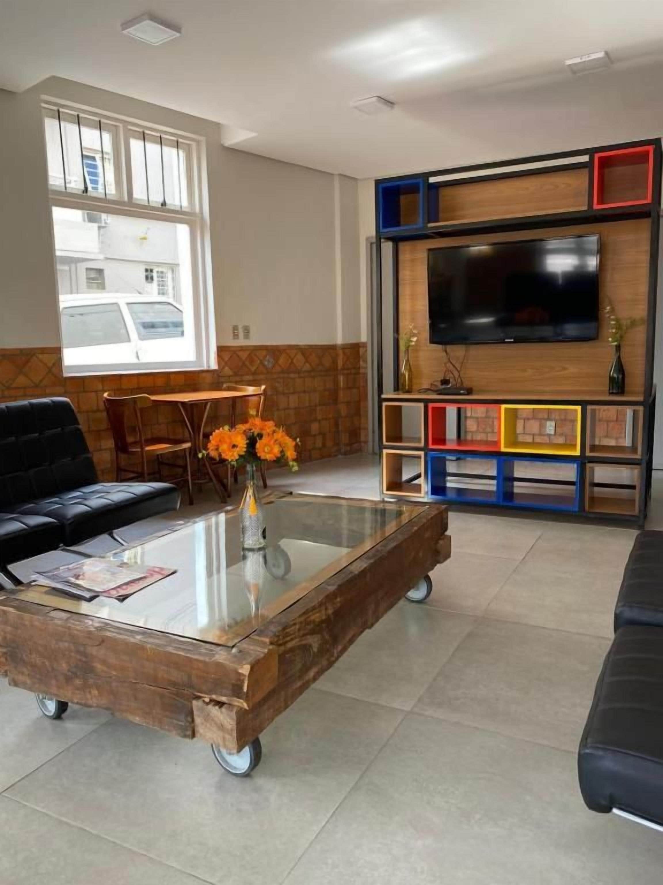 Uma sala de estar com uma mesa de centro de vidro, um sofá preto, uma televisão fixada em uma parede de madeira e uma janela com vista para um edifício.