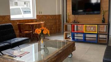 Uma sala de estar com uma mesa de centro de vidro, um sofá preto, uma televisão fixada em uma parede de madeira e uma janela com vista para um edifício.