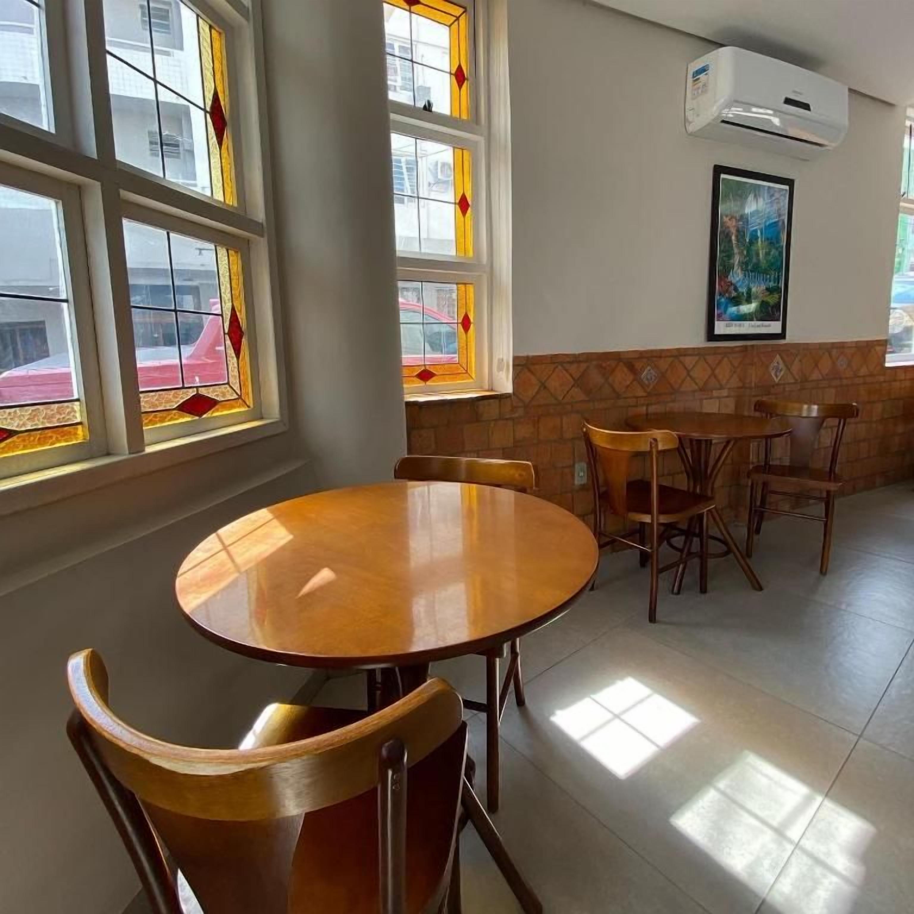 Uma sala de jantar com mesa redonda e cadeiras, janelas amplas com vitrais e um aparelho de ar condicionado na parede.