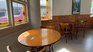 Uma sala de jantar com mesa redonda e cadeiras, janelas amplas com vitrais e um aparelho de ar condicionado na parede.
