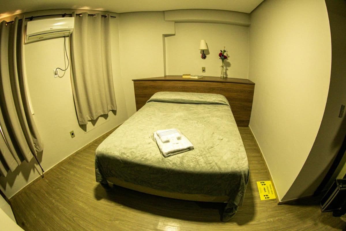 Una habitación de hotel con una cama, un tocador de madera, una mesita de noche con lámpara y un ventanal con cortinas.