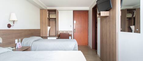 A hotel room with two beds, a desk, a chair, a TV mounted on the wall, and a wooden wardrobe.