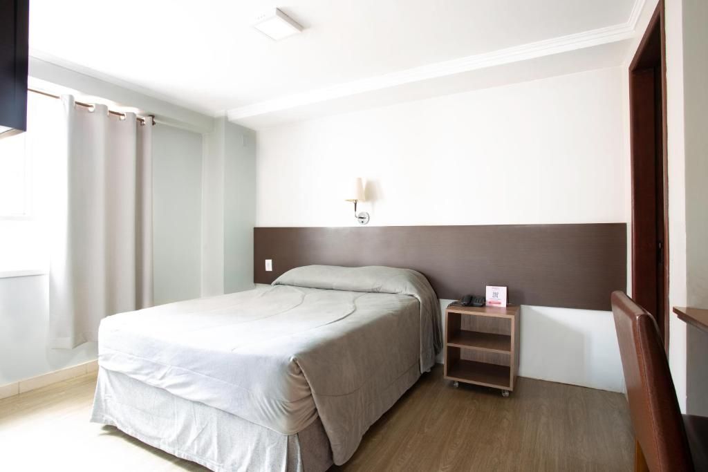 A hotel room with a bed, a nightstand, a wall-mounted lamp, and a window with curtains.