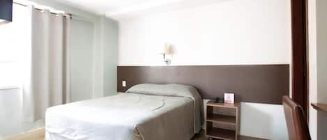 A hotel room with a bed, a nightstand, a wall-mounted lamp, and a window with curtains.