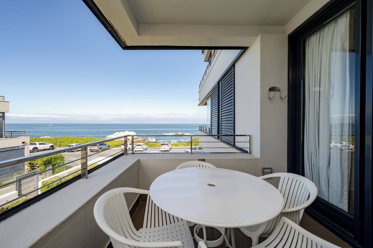 A balcony with a white table and chairs, overlooking a coastal view with parked cars and a clear sky.