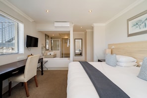 Une chambre d’hôtel avec un lit, un bureau, une chaise, une télévision et une salle de bain visible à travers une porte ouverte.