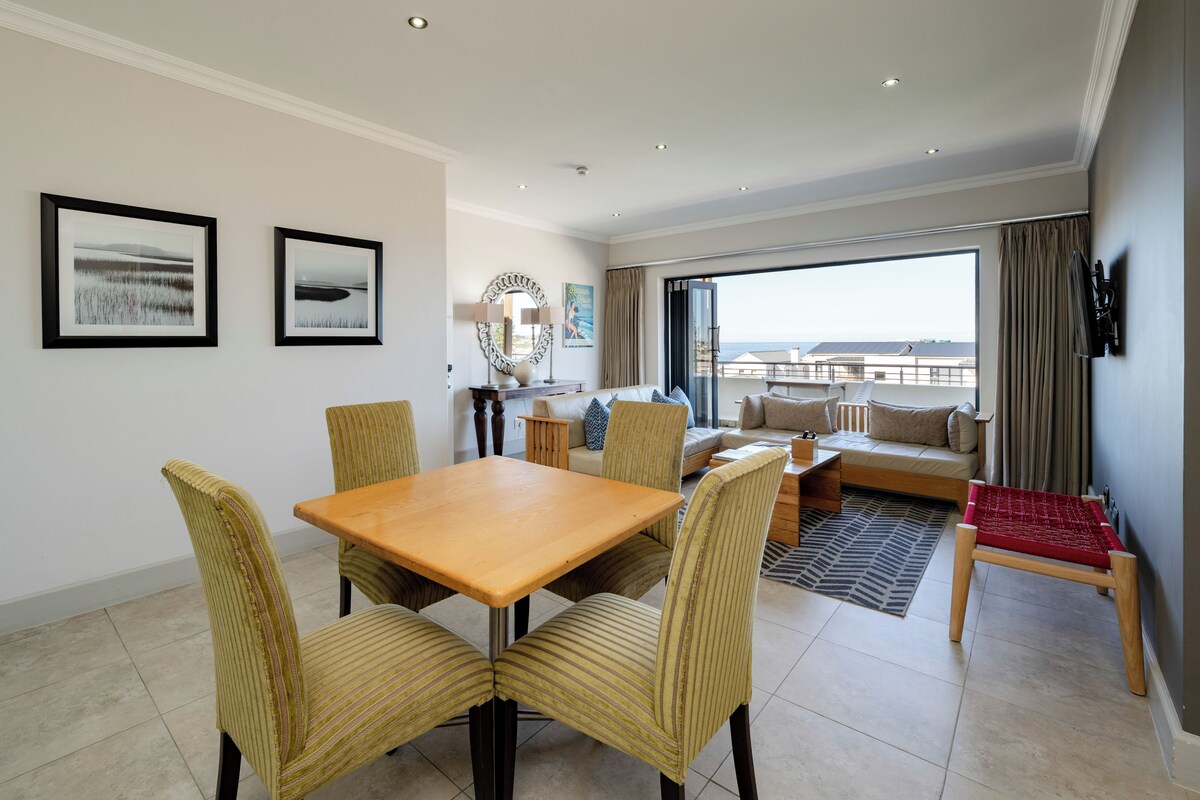 A hotel room with a dining area, a sofa, a TV, and a view of the sea.