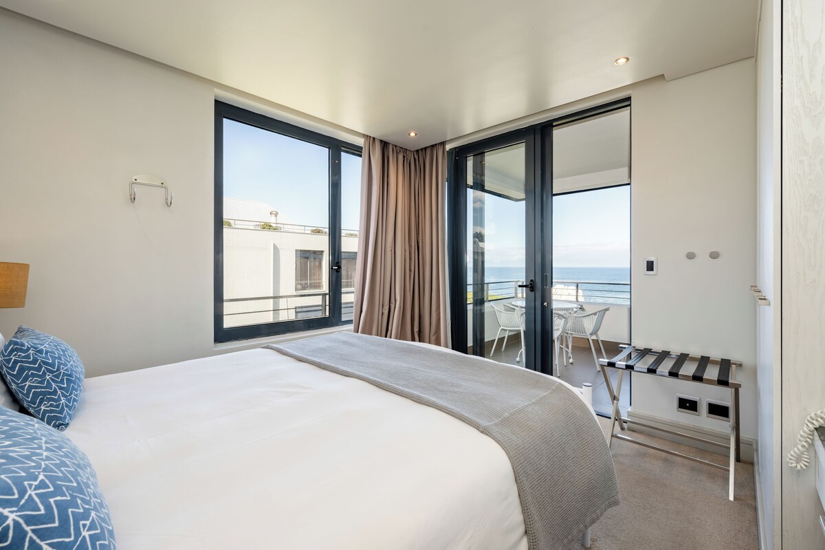 A modern hotel room with a large bed, a balcony with a table and chairs, and a view of the sea.