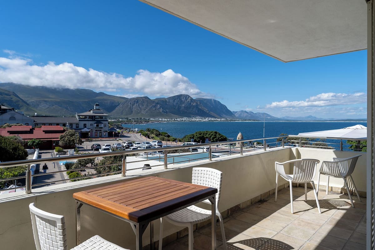 A balcony with a wooden table and white chairs, overlooking a coastal town with mountains in the background.
