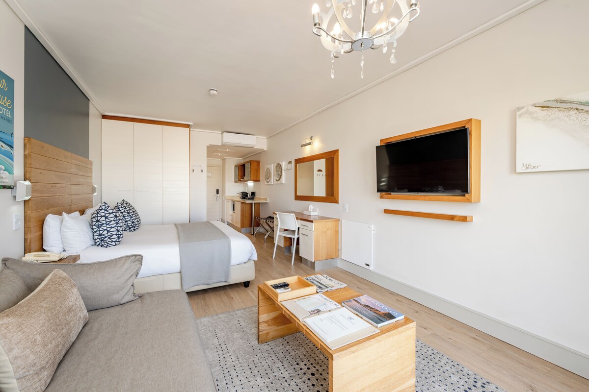 A modern hotel room with a bed, a desk, a chair, a sofa, a TV, and a chandelier.