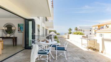 An outdoor patio with a round table and chairs, a grill, and a view of the sea.