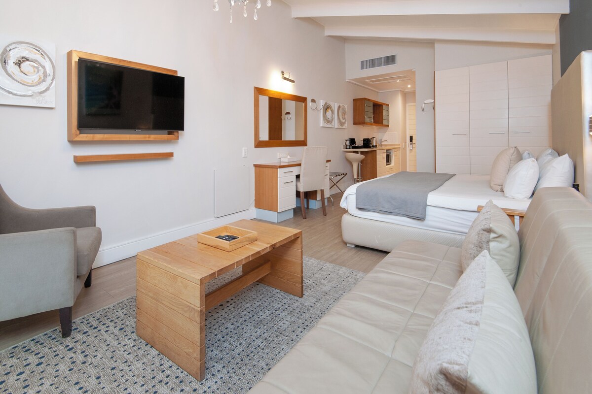 Una sala de estar moderna y bien iluminada, con un sofá, una mesa de centro y un televisor montado en la pared. Cuenta con una pequeña cocina con armarios y un lavamanos.