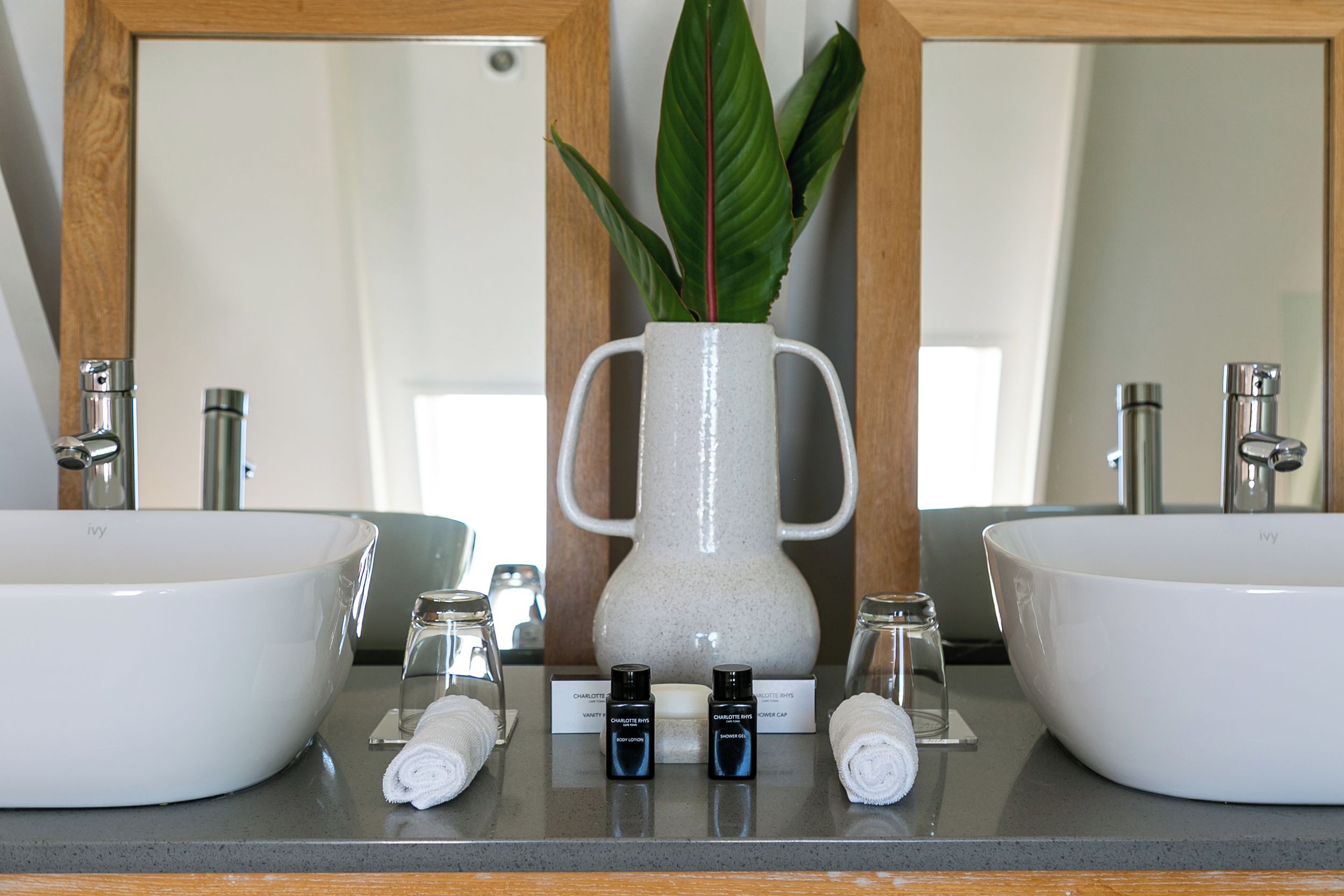 A bathroom with two sinks, a mirror, a vase with a plant, and toiletries.