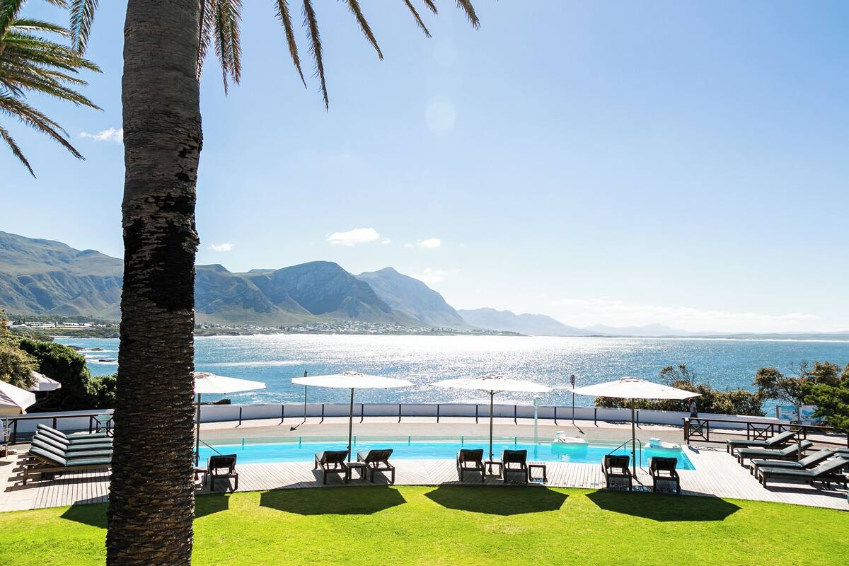 A poolside area with lounge chairs and umbrellas, overlooking a beach and mountains.