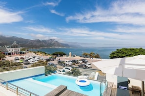 A rooftop pool with a view of the ocean and mountains, surrounded by parked cars and a building with a dome. - Harbour House Hotel (Hermanus)