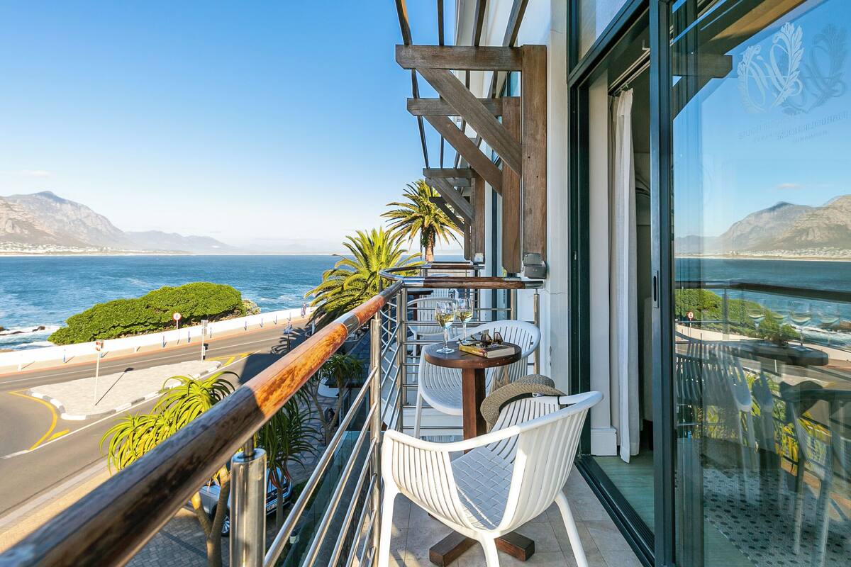 A balcony with a view of the ocean, white chairs, and a table with a glass of wine.