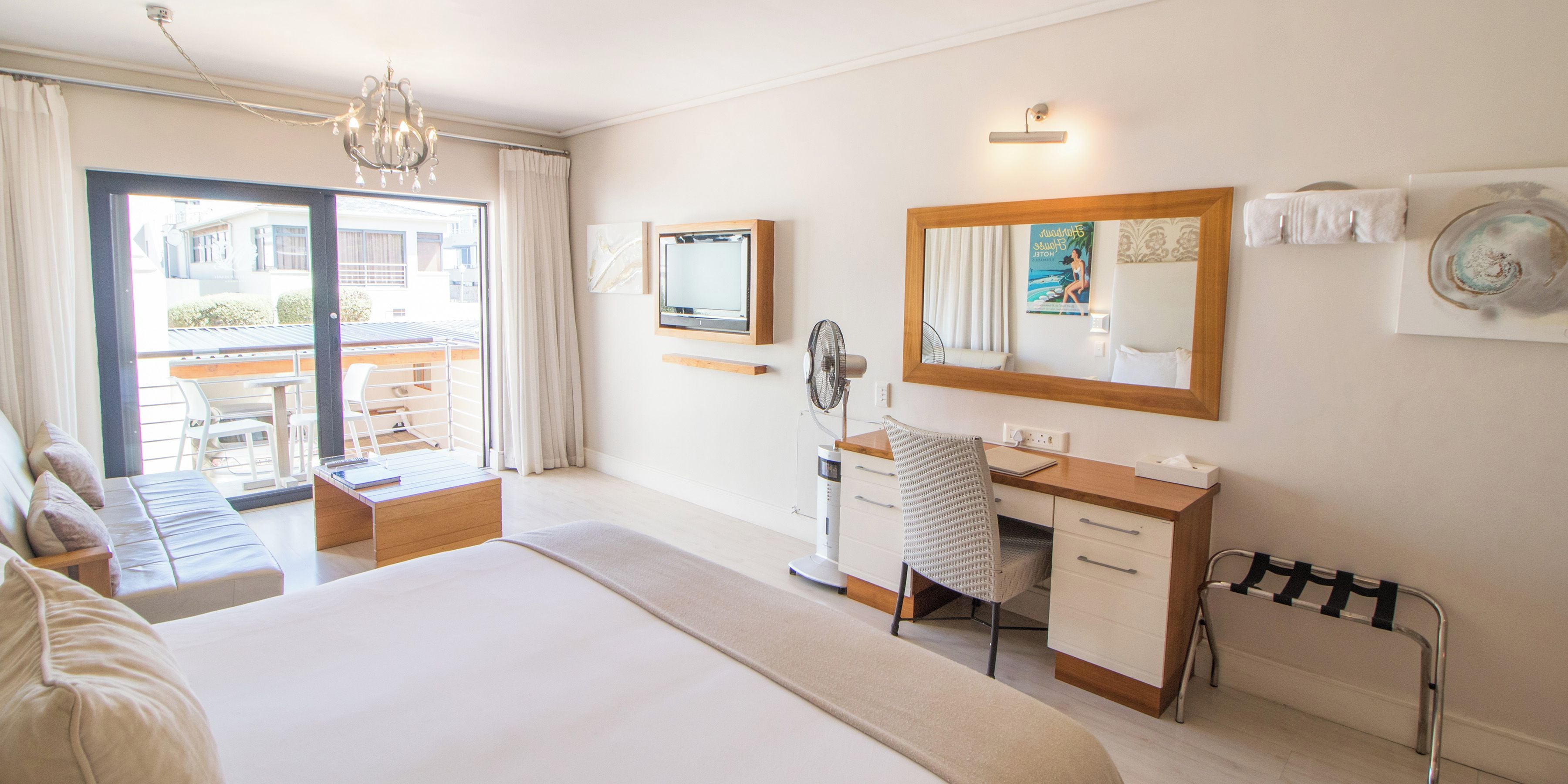 Ein Hotelzimmer mit einem Bett, einem Schreibtisch, einem Stuhl, einem Standventilator, einem Fenster mit Vorhängen und einem Balkon mit Gartenmöbeln.