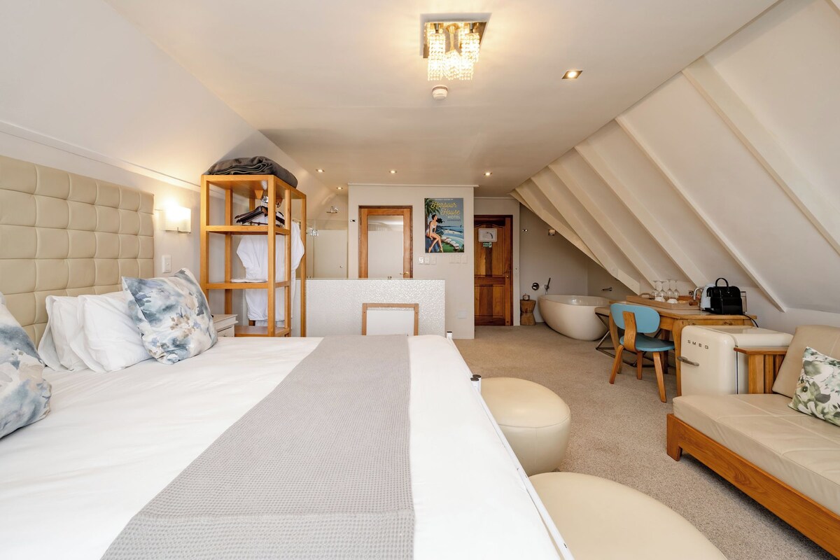 A hotel room with a large bed, a wooden wardrobe, a sitting area with a table and chairs, a bathtub, and a kitchenette.