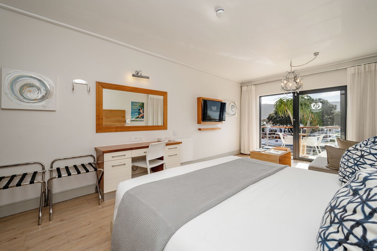 A hotel room with a bed, desk, chair, and a large window with a view of a balcony.