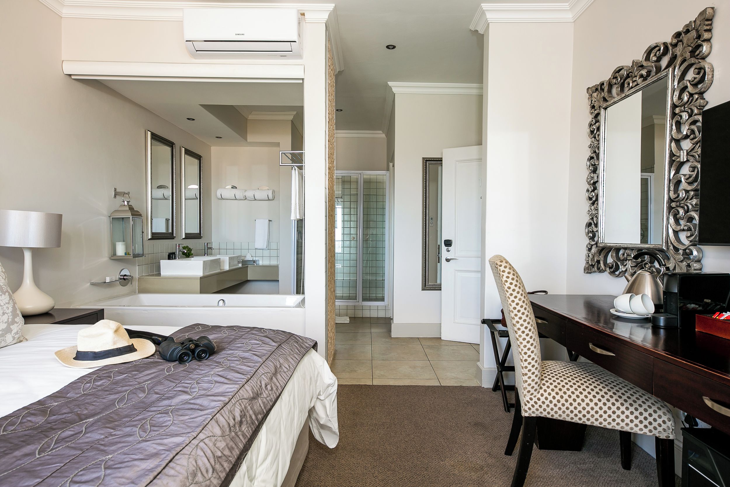 A hotel room with a bed, a desk, a chair, a large mirror, and a bathroom visible through an open door.