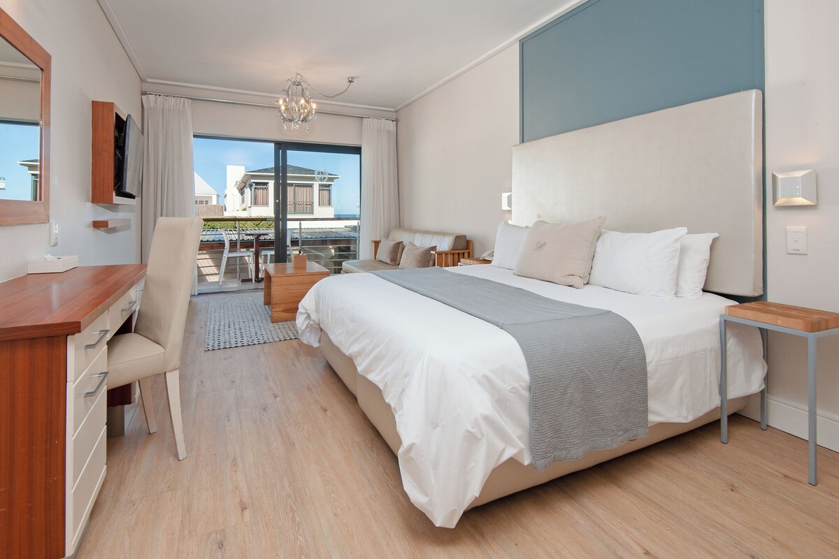 A hotel room with a large bed, a desk, a chair, a small table, and a view of the outside.