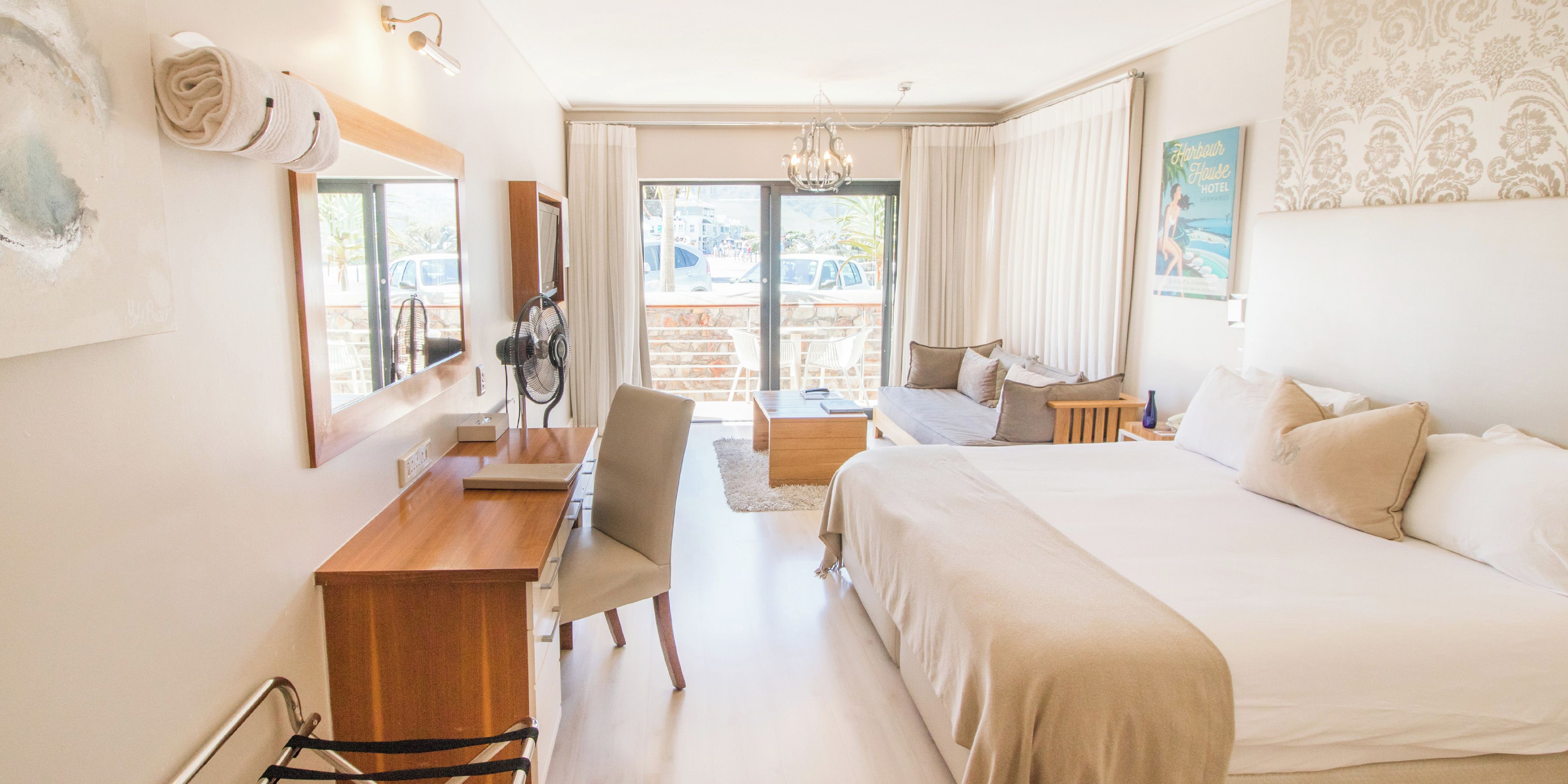 A hotel room with a bed, a desk, a chair, a fan, and a view of the outside.