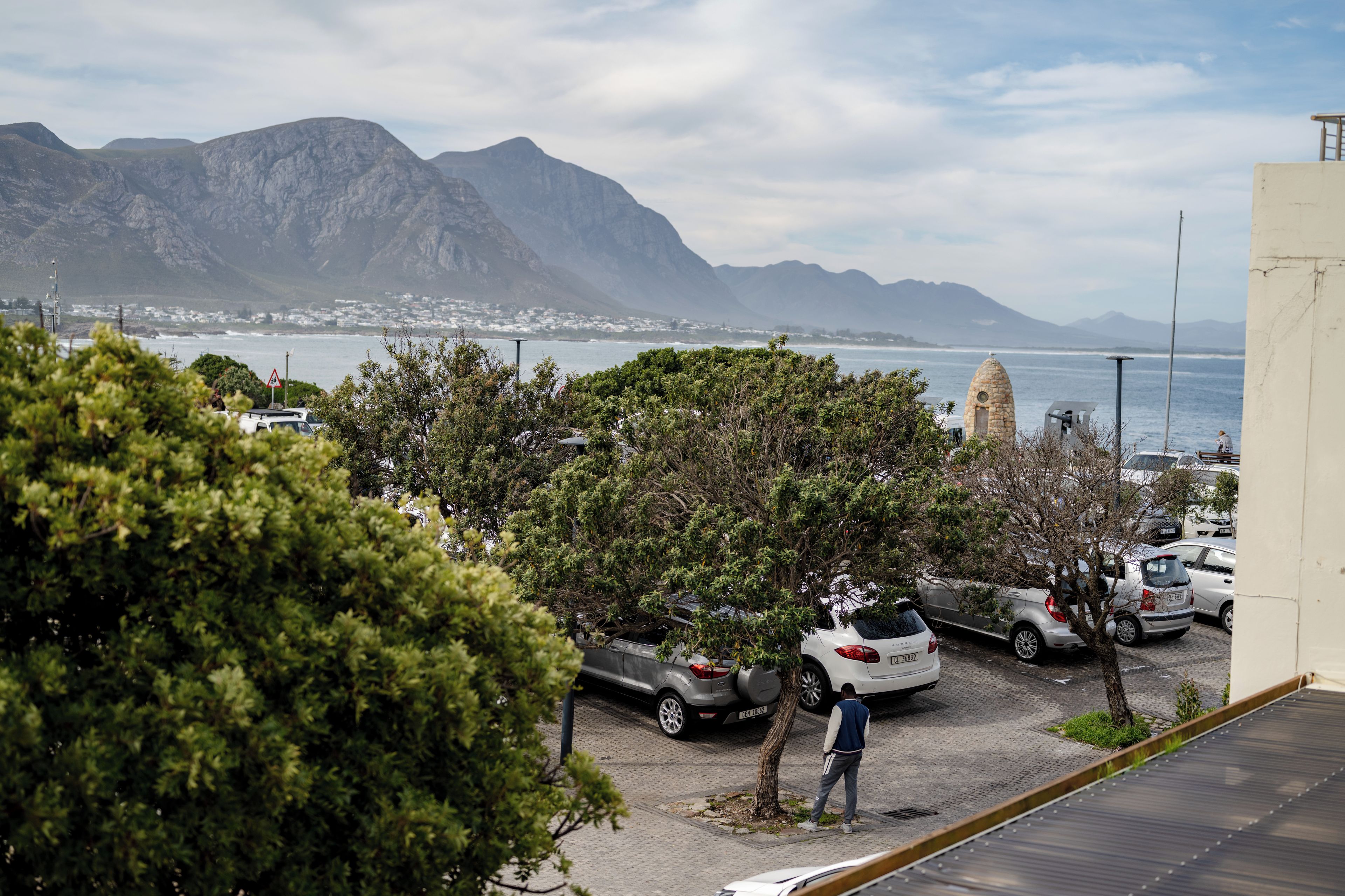 Un estacionamiento con coches, una persona caminando y vistas a las montañas y al mar.