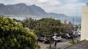A parking area with cars, a person walking, and a view of mountains and the sea.