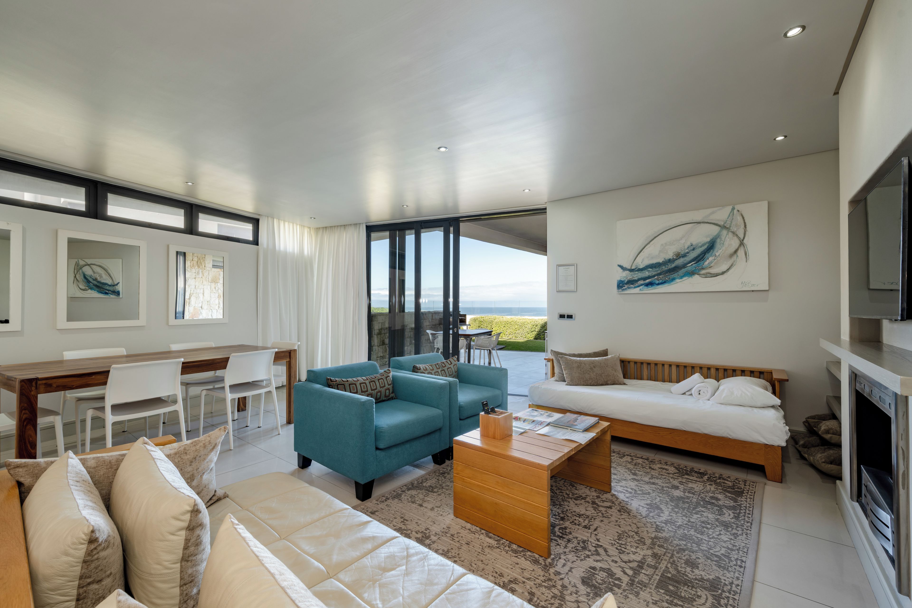 A modern living room with a sofa, coffee table, and chairs. There is a dining area with a table and chairs. A large window offers a view of the outdoors.