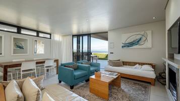 A modern living room with a sofa, coffee table, and chairs. There is a dining area with a table and chairs. A large window offers a view of the outdoors.