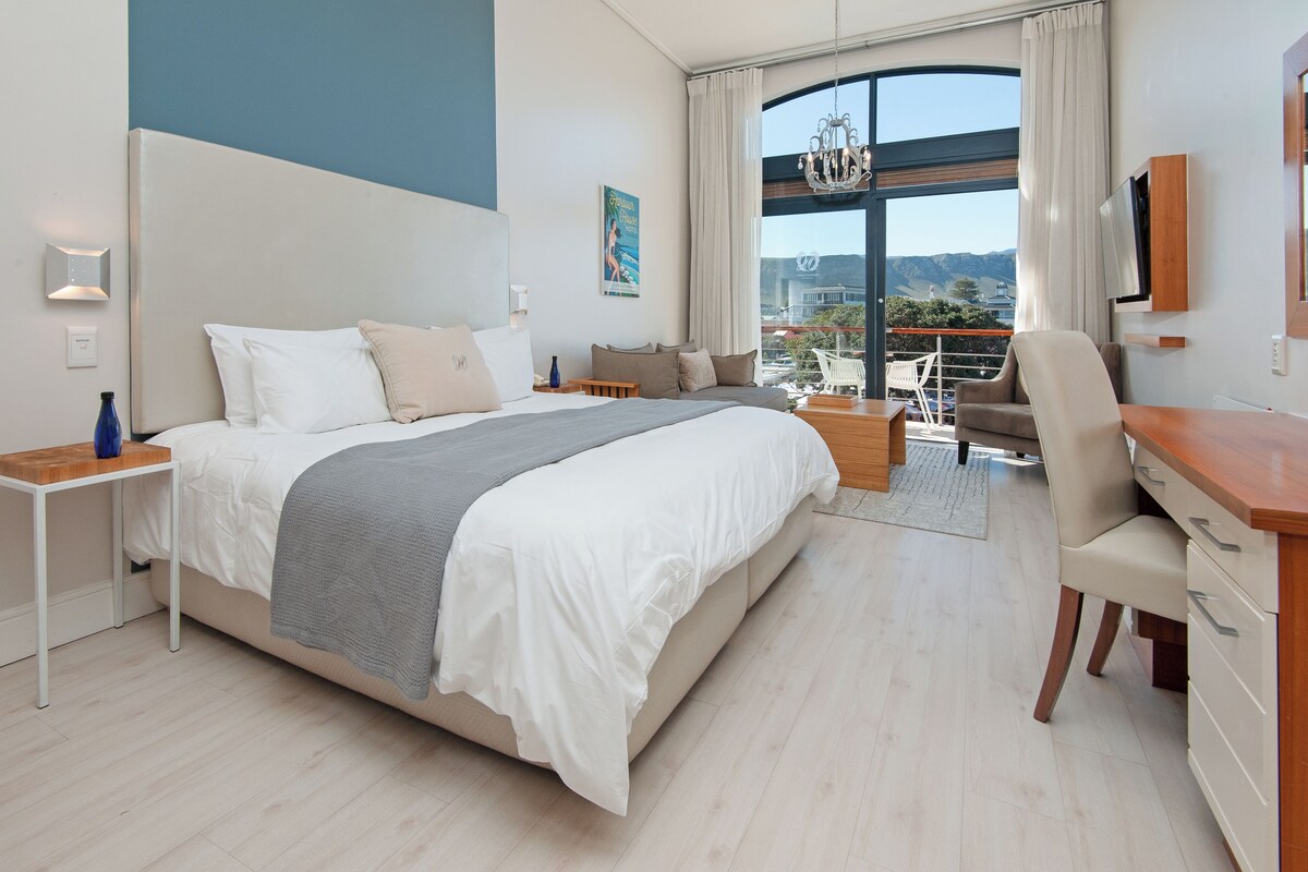 A modern hotel room with a large bed, a desk, and a balcony with a view.