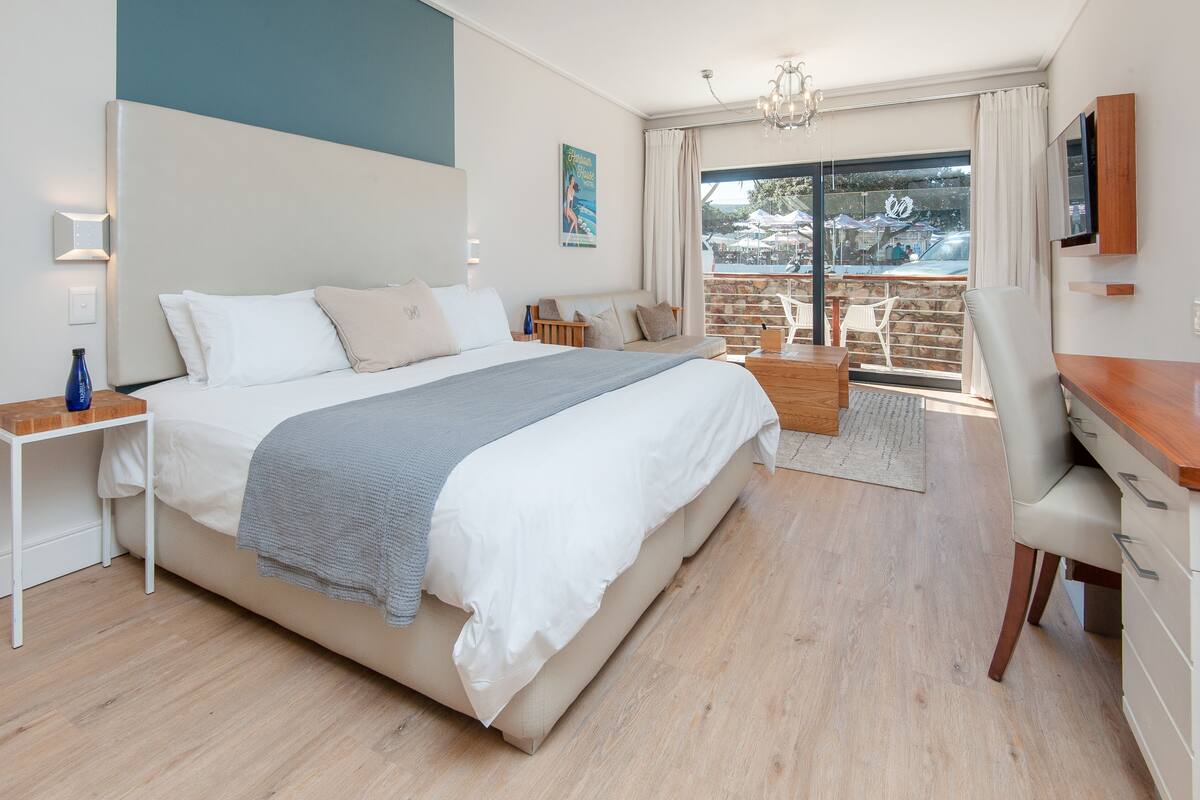 A modern bedroom with a large bed, a desk, and a balcony with outdoor seating.