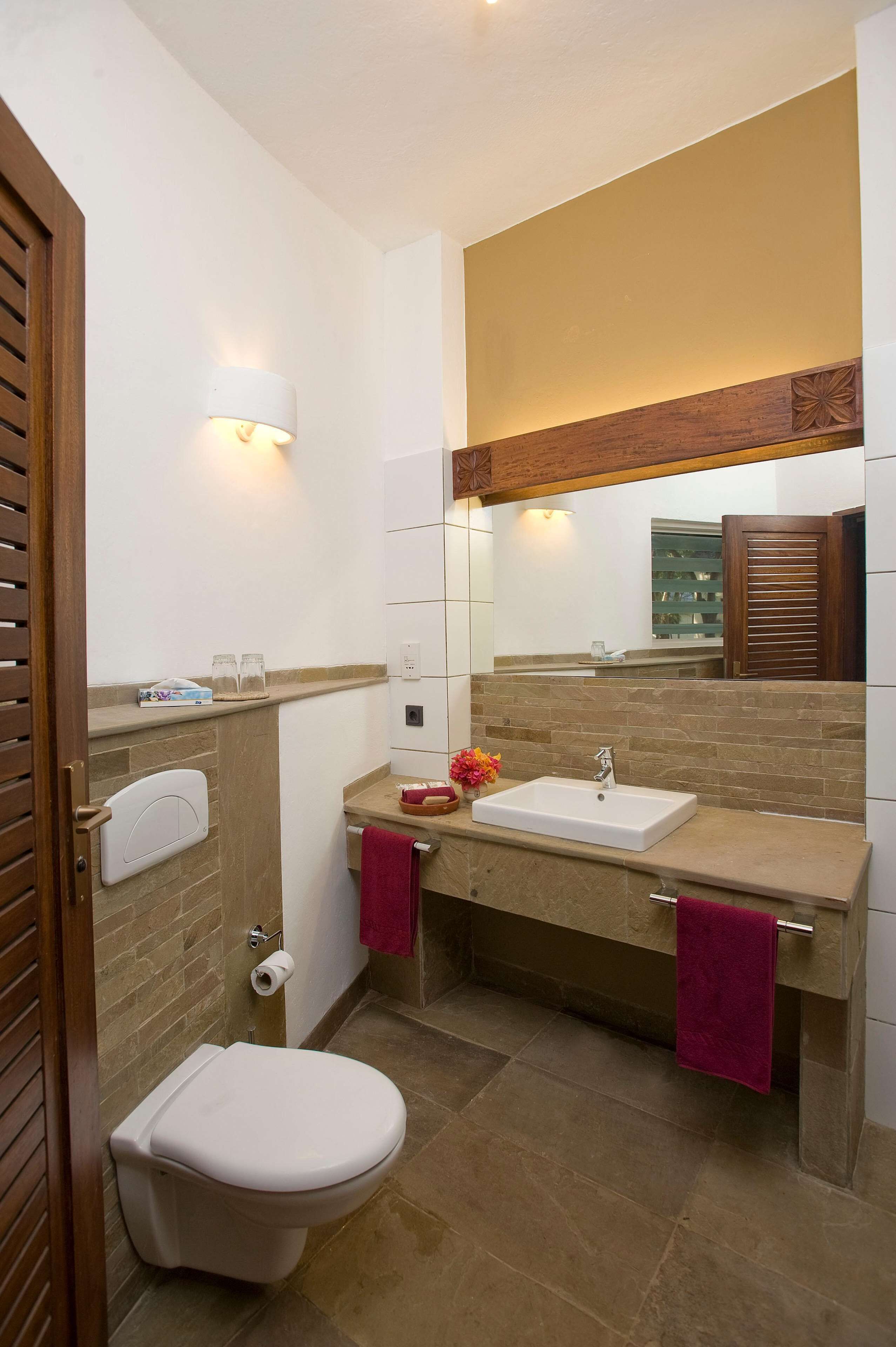 Une salle de bain avec des toilettes blanches, deux lavabos dotés de plans de travail beiges, un miroir au-dessus des lavabos et une porte en bois.