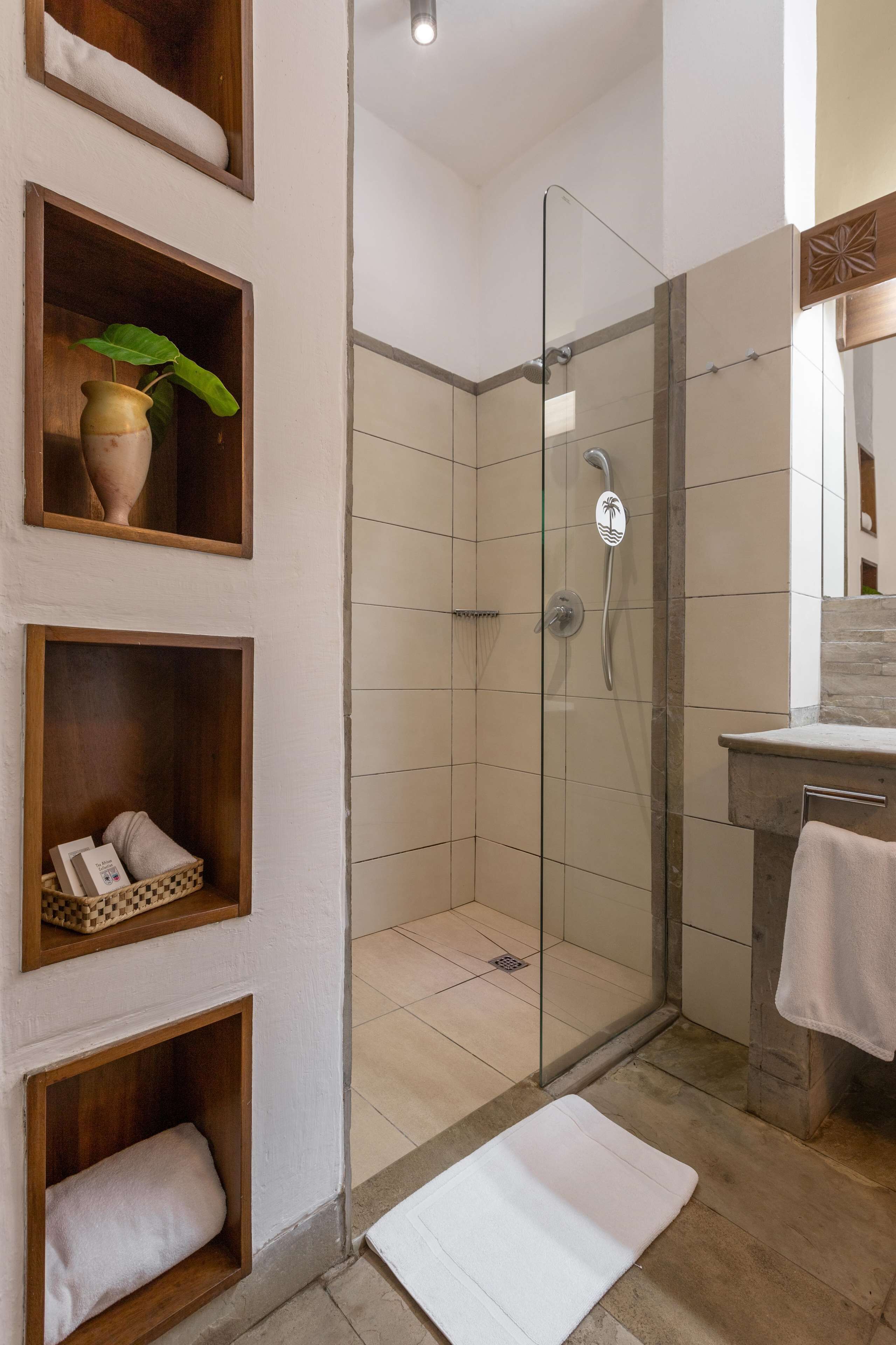 Une salle de bain moderne avec une douche à parois de verre, des étagères en bois où sont posées des serviettes et une plante, et un tapis de bain blanc sur le sol.