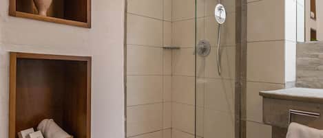 Un baño moderno con ducha envuelta en cristal, repisas de madera con toallas y una planta, y un tapete de baño blanco en el piso.