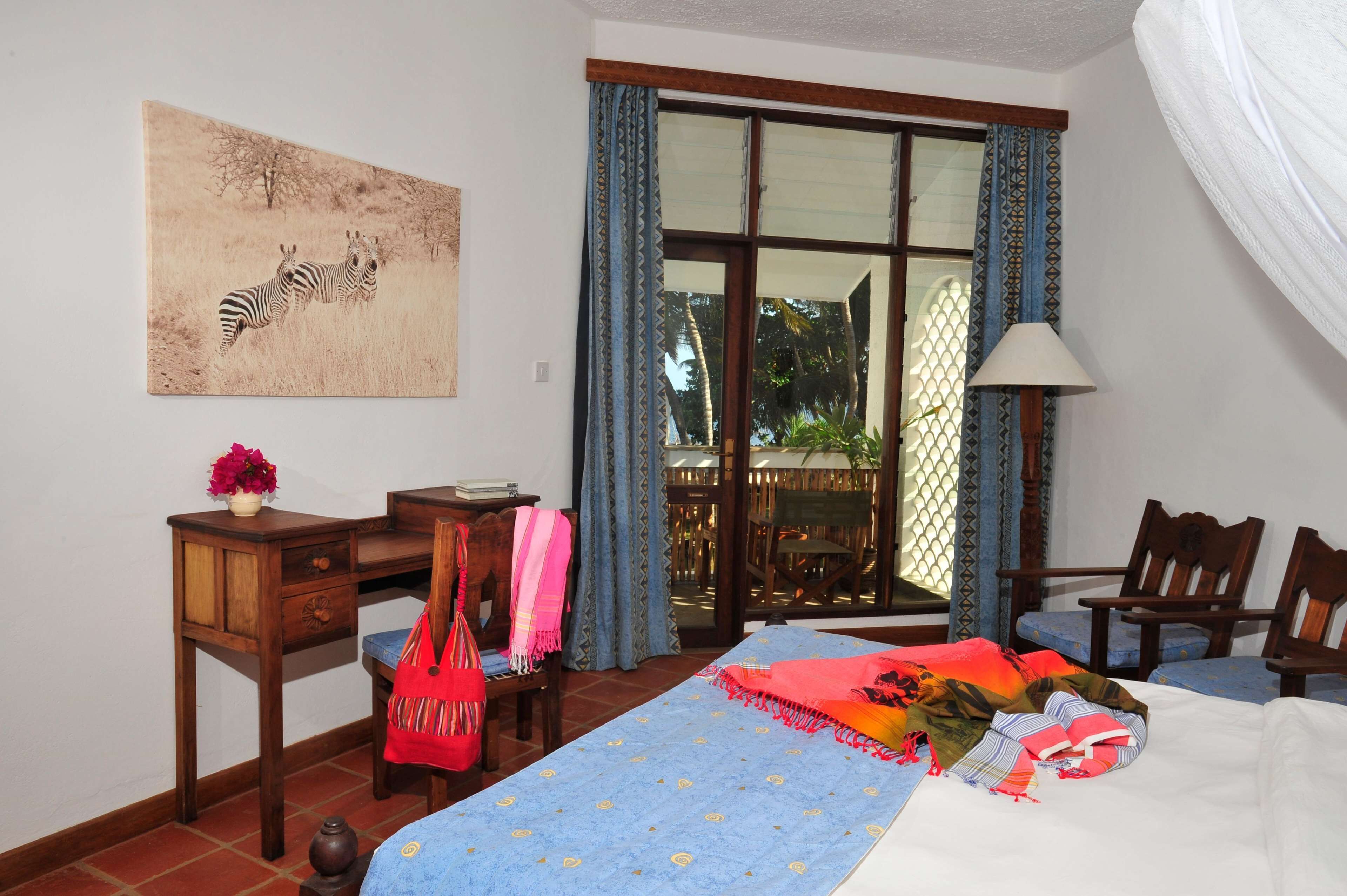 Une chambre à coucher comprenant un lit, un bureau, une chaise, une fenêtre avec des rideaux, une lampe et un tableau représentant des zèbres.