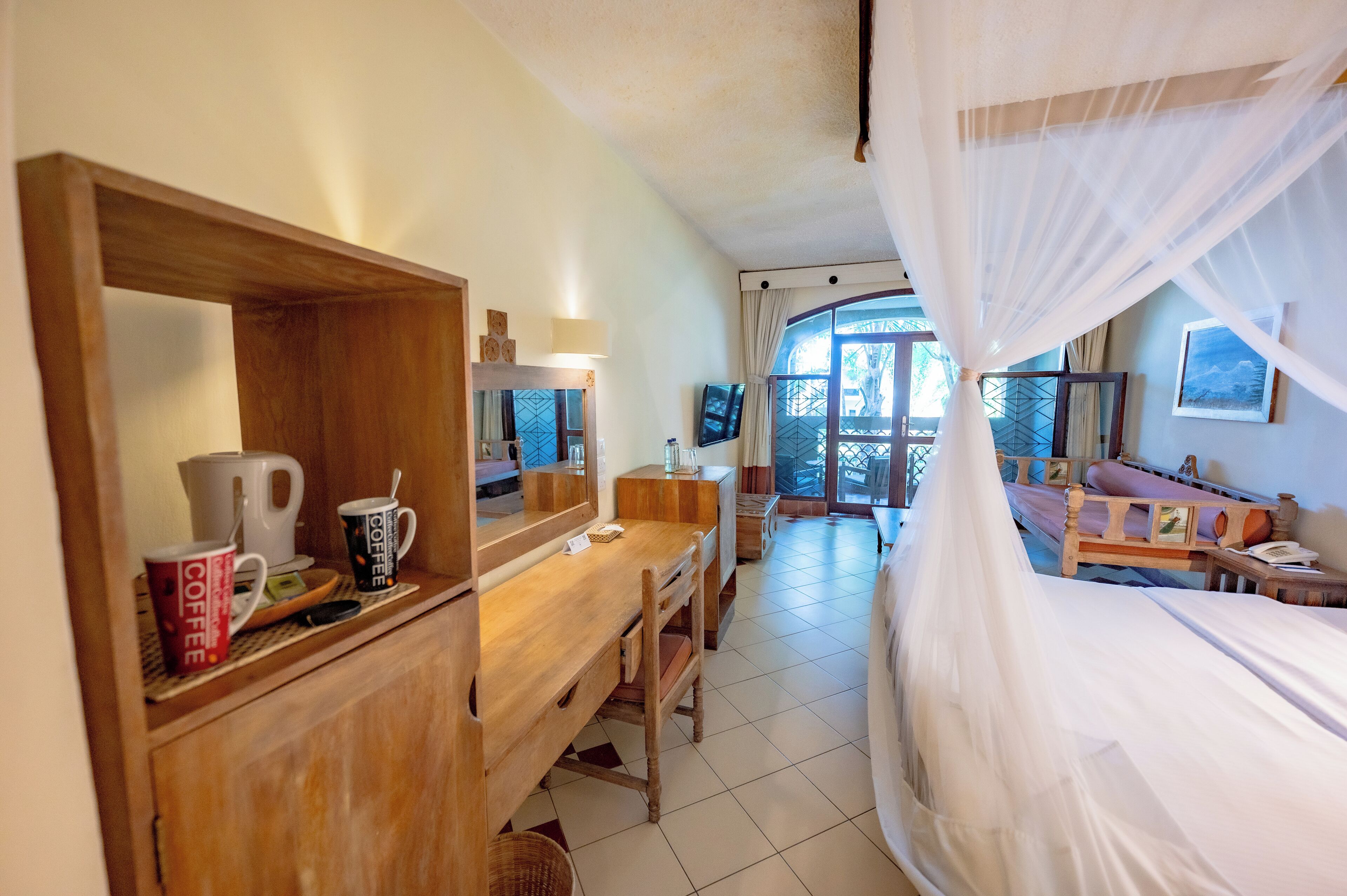 A room with a wooden desk, a bed with white curtains, a television, and a view of the outdoors.