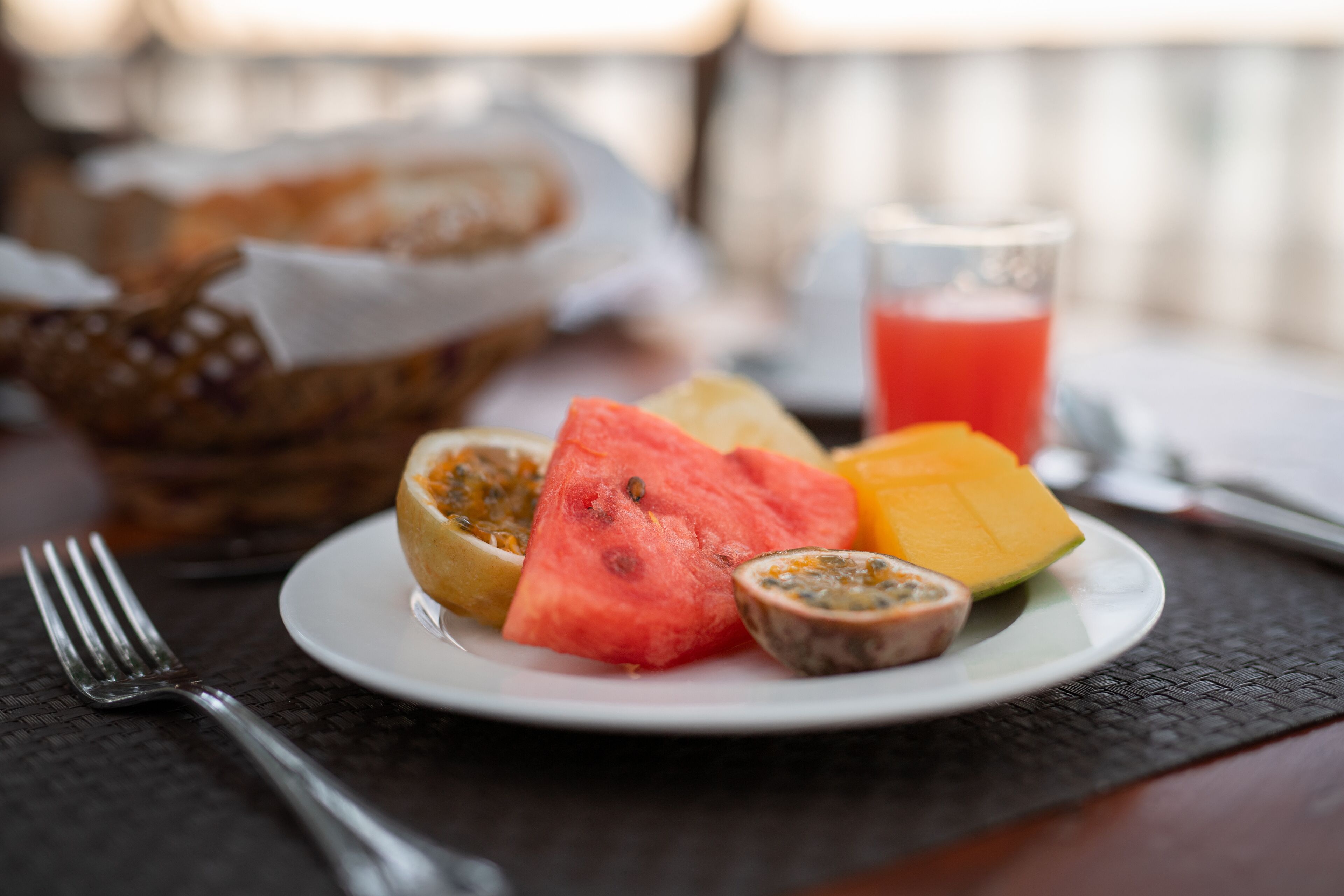 A plate with sliced watermelon, mango, passion fruit, and a glass of red juice on a table with a fork and a basket of bread in the background.