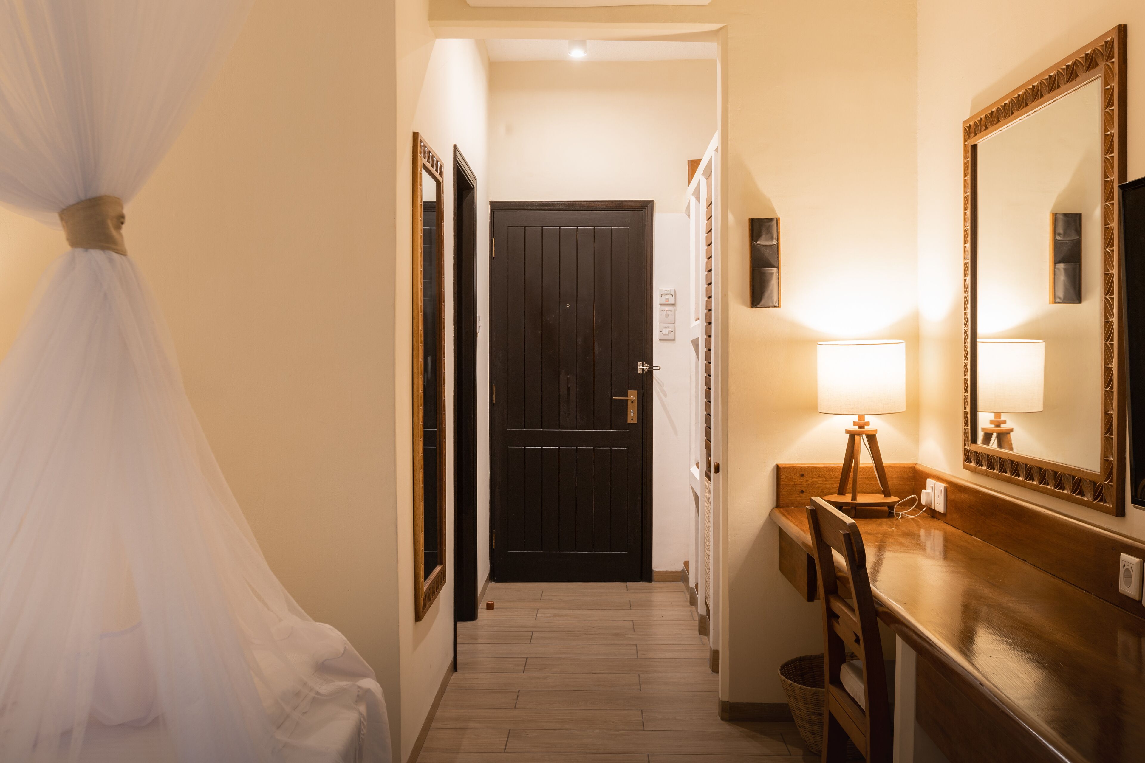 A hallway with a dark door, a wooden bench, a mirror, and a bedside table with a lamp.