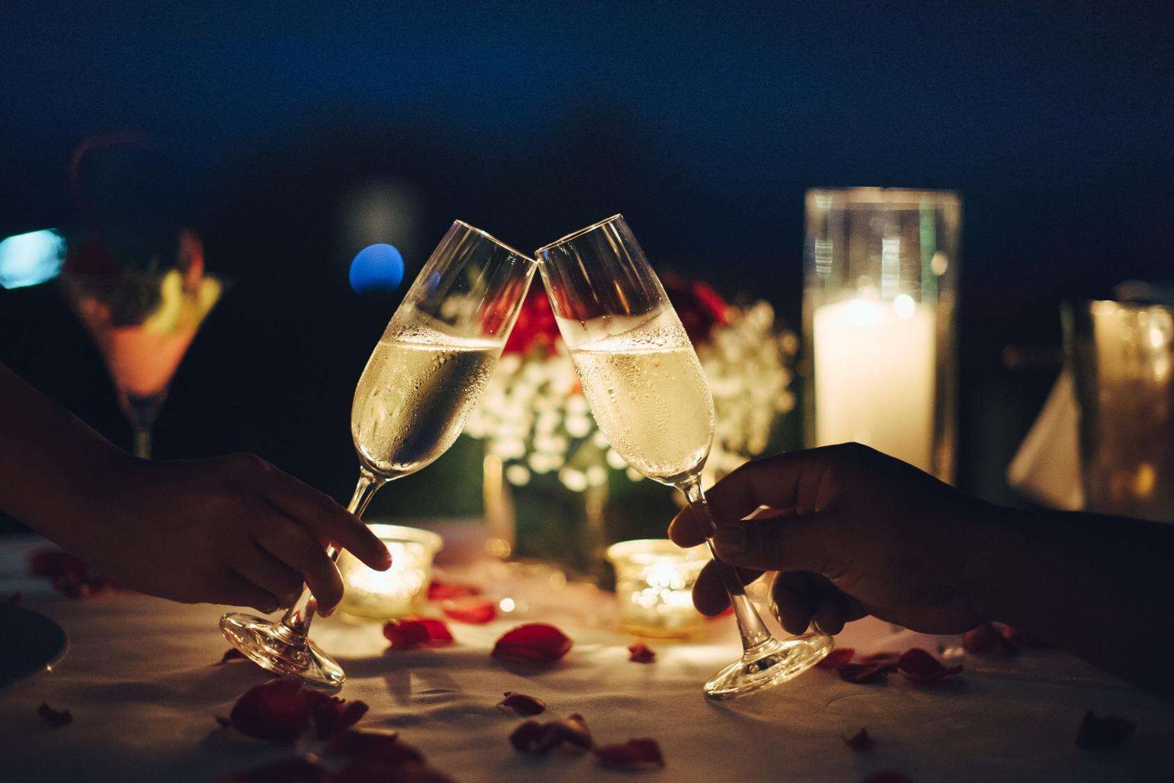 Two hands holding champagne flutes, a candle, and rose petals on a table.