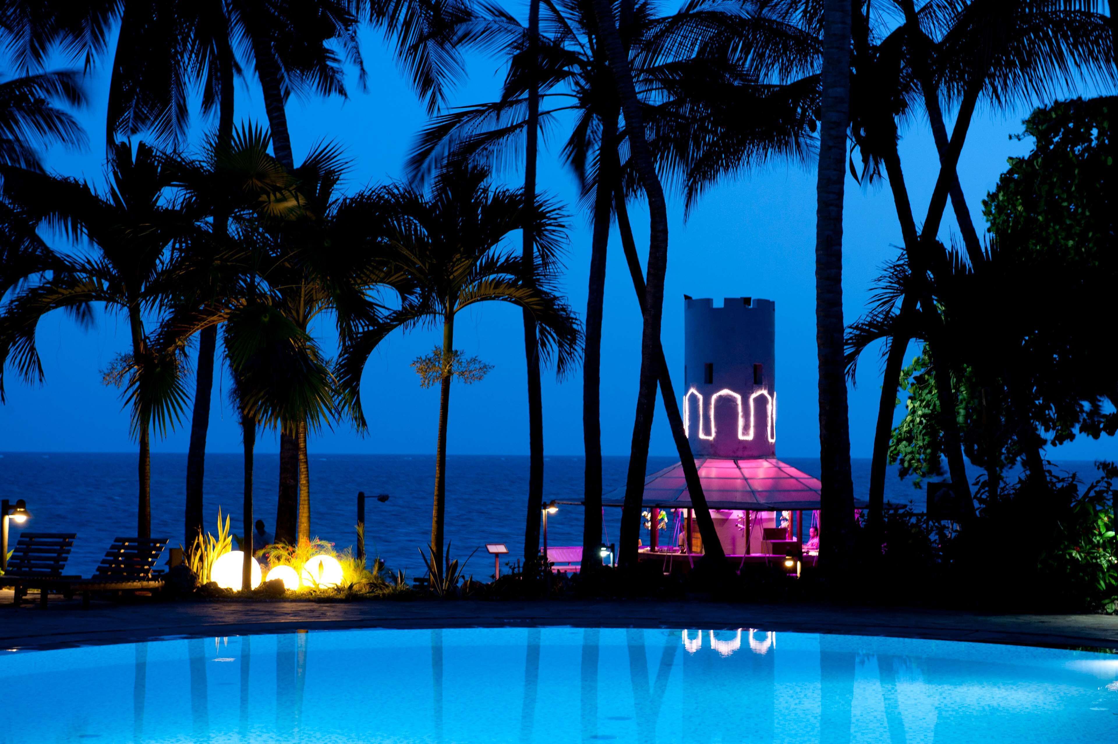 A poolside view with palm trees, a beach, and a building with a neon sign at dusk.