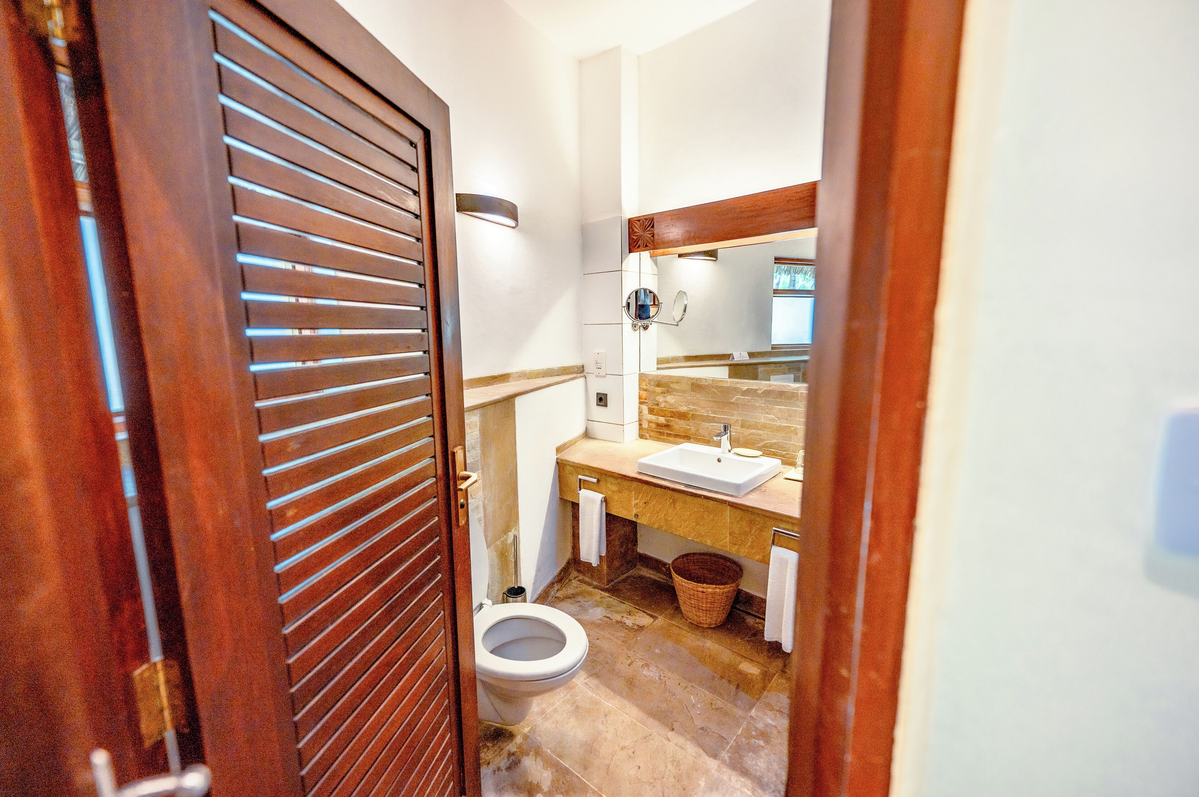 Une petite salle de bain avec une porte en bois, des toilettes blanches, un lavabo avec un miroir et un sol carrelé.