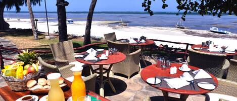A beachside restaurant with outdoor seating, tables set with plates, glasses, and a fruit basket.