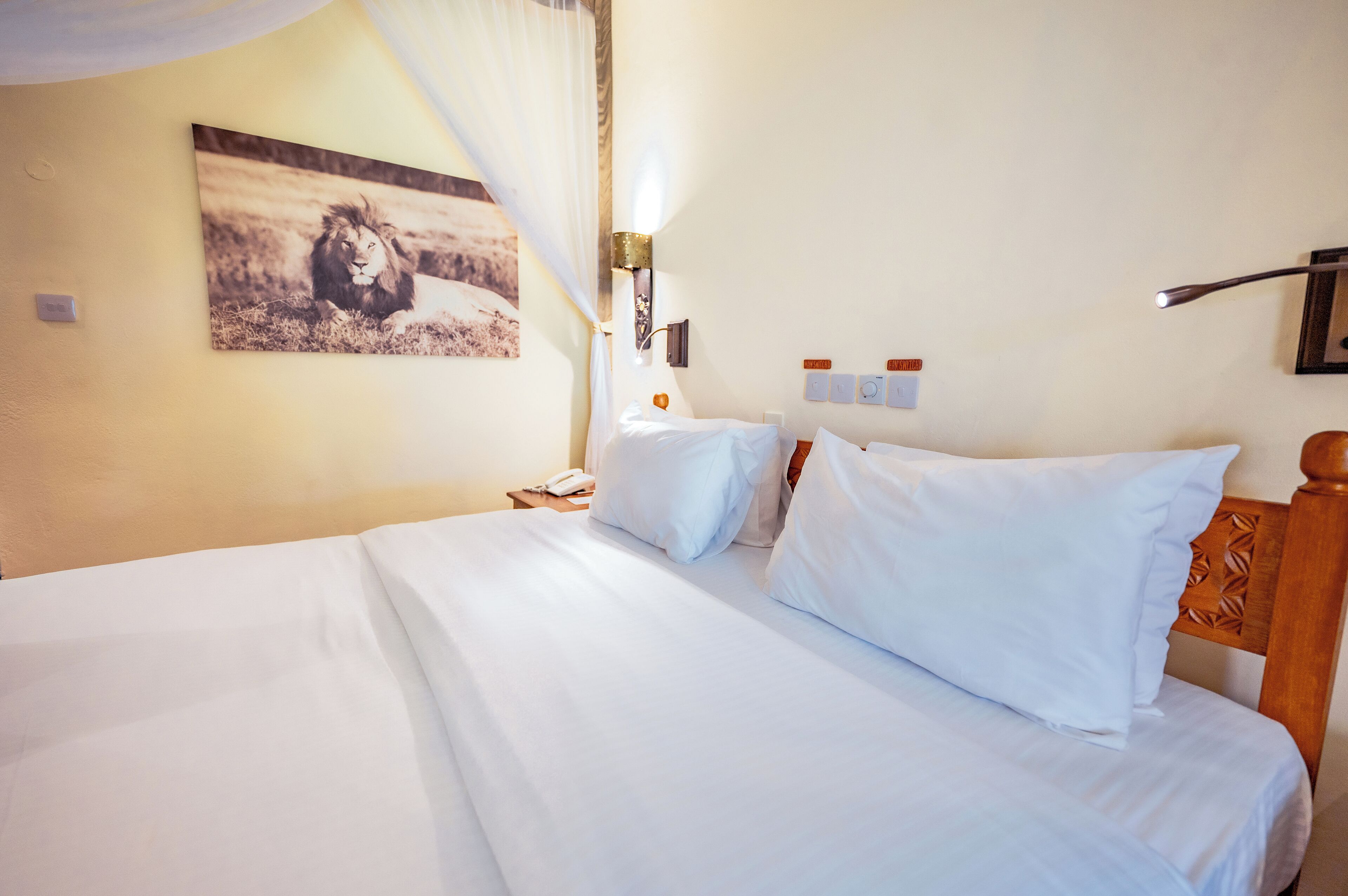 A hotel room with a large bed, a bedside table with a lamp, a wall-mounted phone, and a picture of a lion.