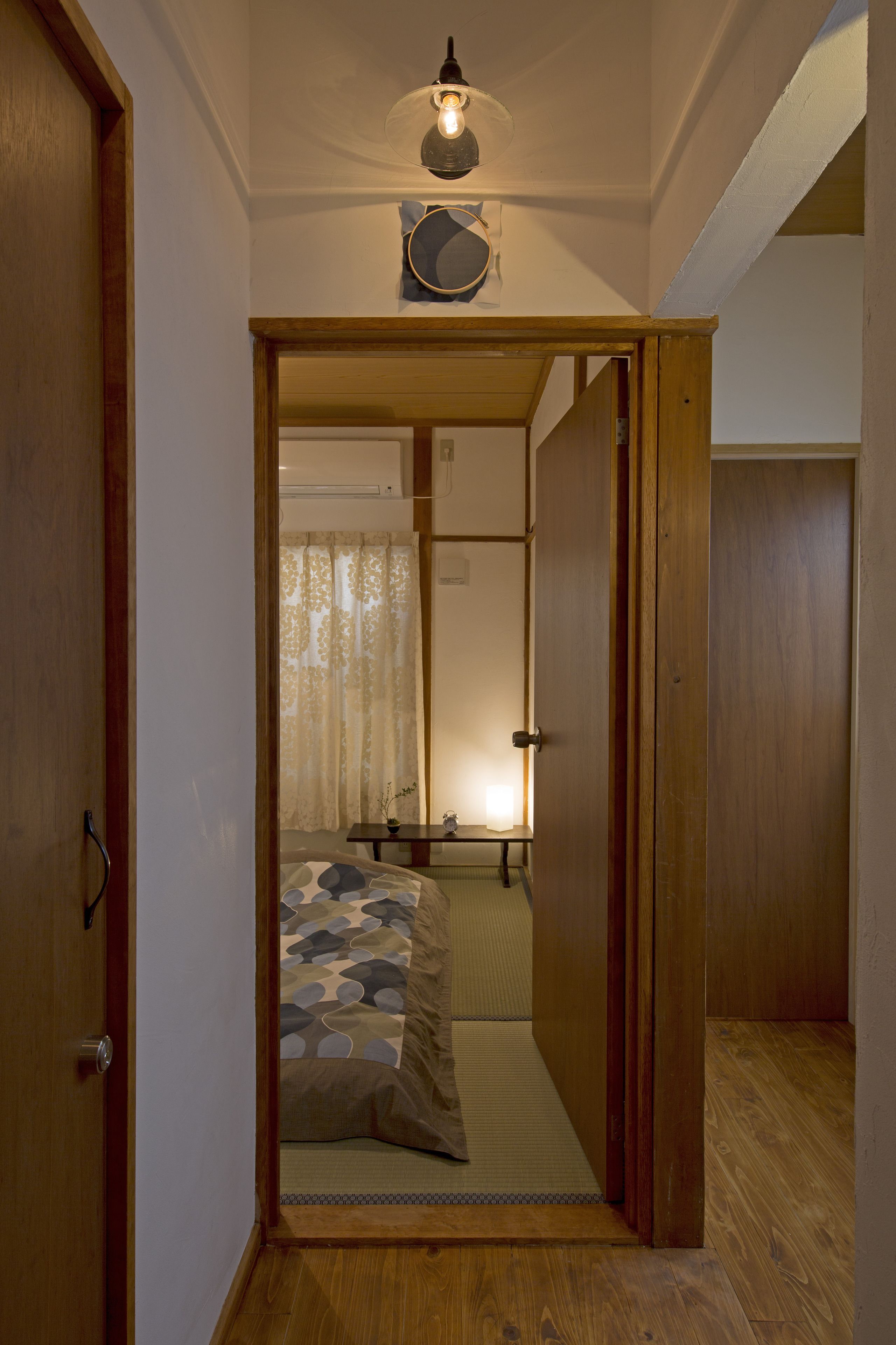 A narrow hallway leading to a bedroom with a bed, a bedside table, and a lamp.