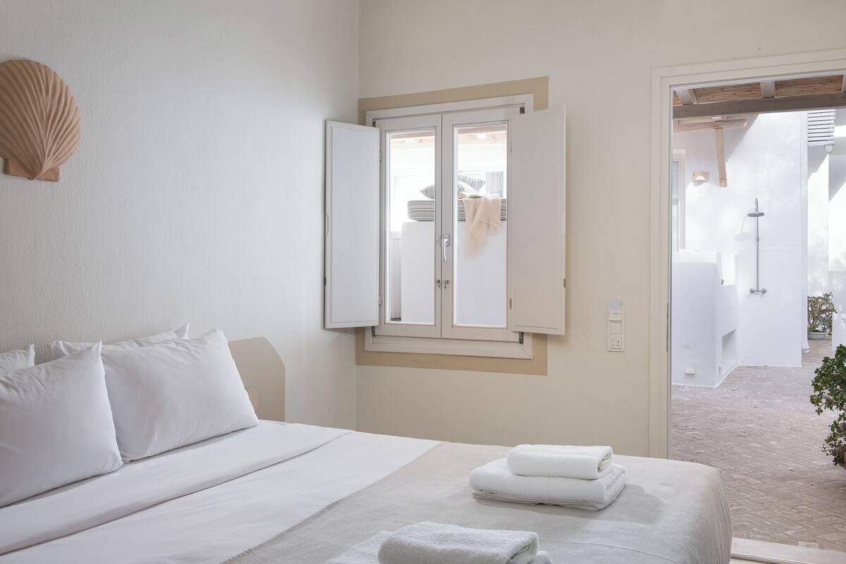 Un dormitorio con una cama, una ventana con contraventanas abiertas y vistas al baño.